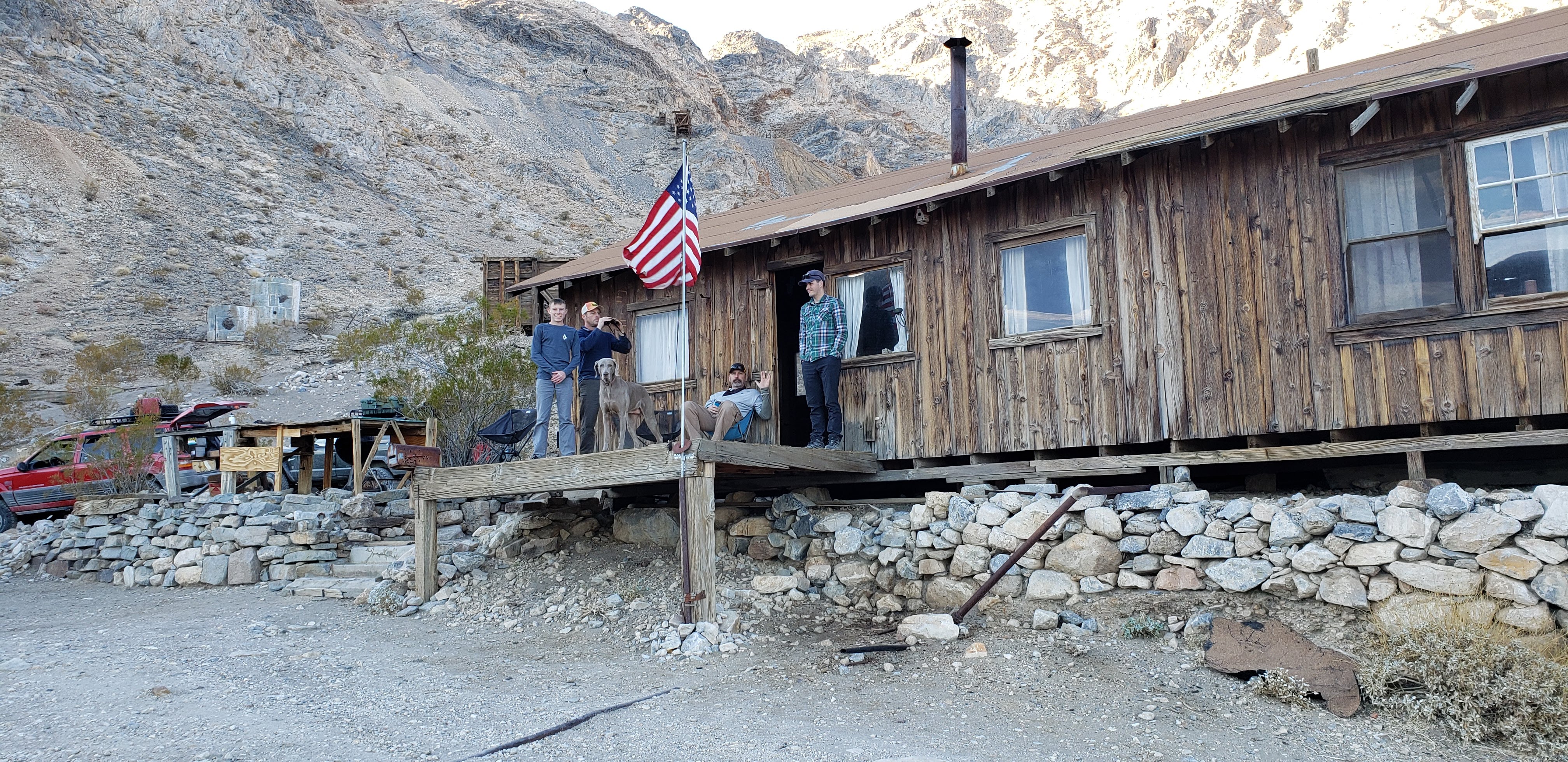 Guys and a dog standing on a wooden porch in front of a wooden cabin.