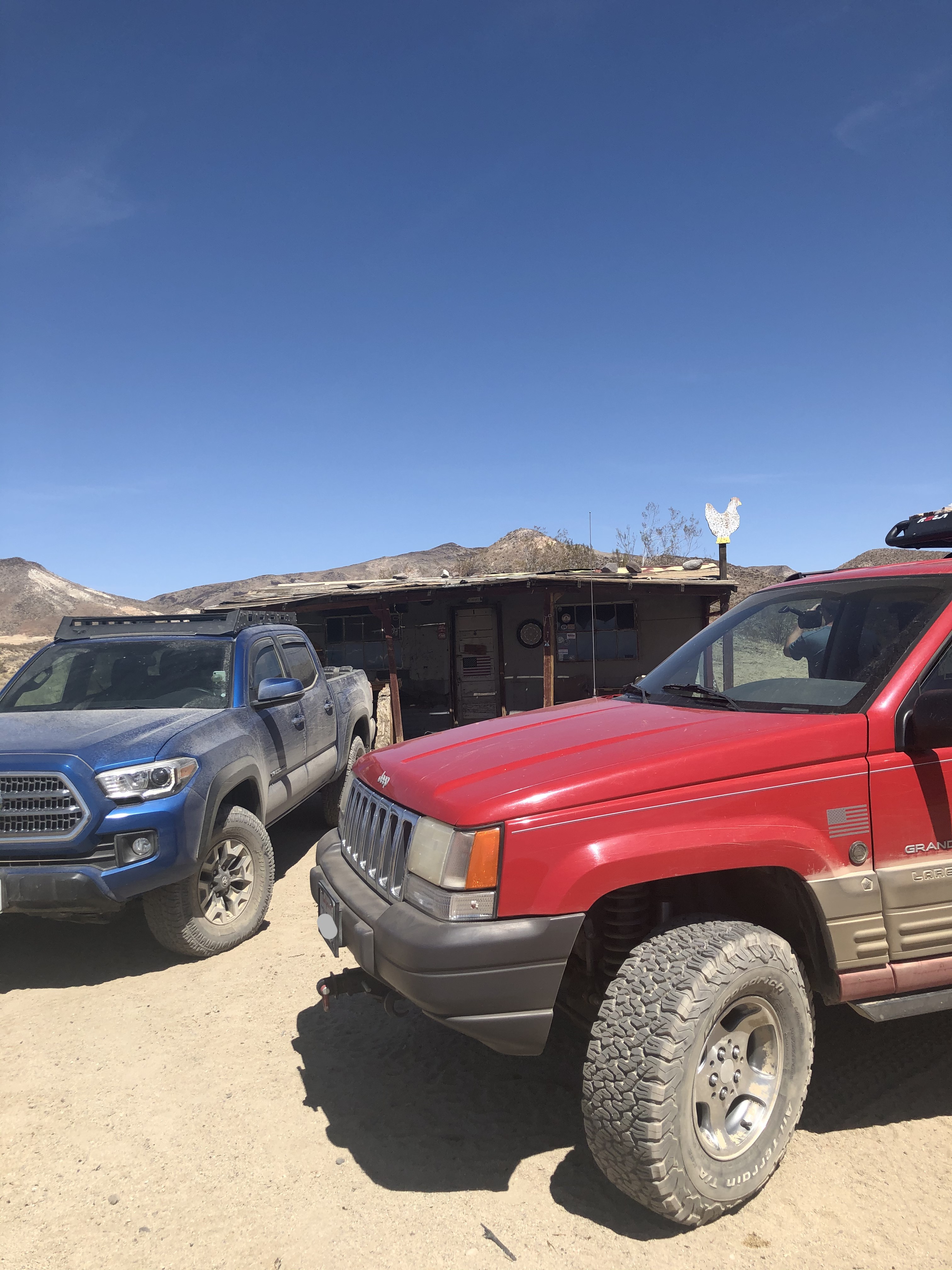 A blue Tacoma and a red ZJ parked in front of an old cabin in the desert.